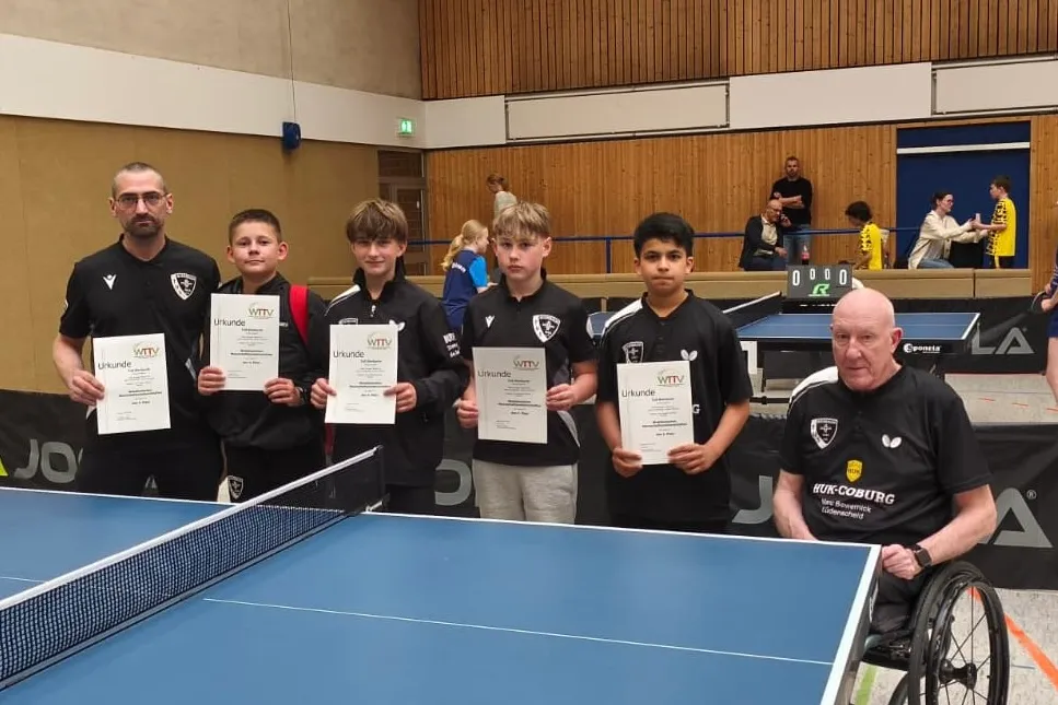 Fünf Jugendliche und ein Mann im Rollstuhl stehen in einer Sporthalle und halten Urkunden. Im Hintergrund ist eine Tischtennisplatte und eine Anzeigetafel zu sehen.