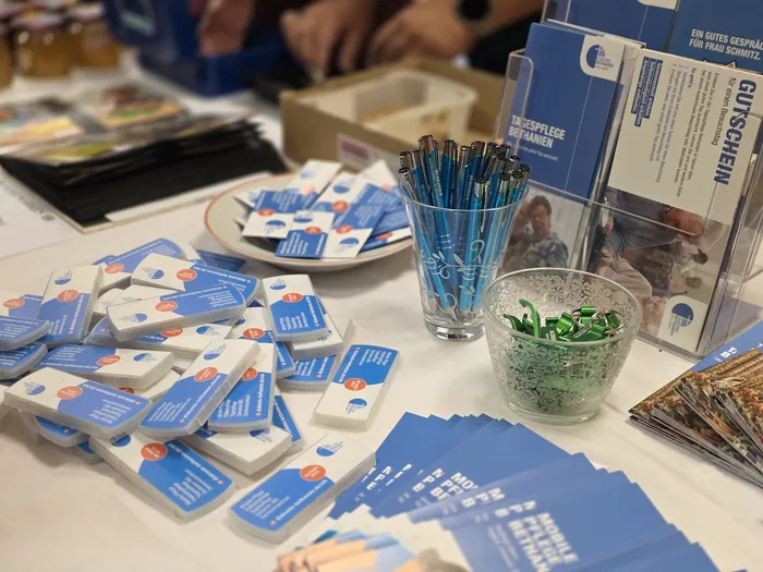 Ein Tisch mit Informationsmaterialien und Werbeartikeln der Tagespflege Bethanien steht im Mittelpunkt. Darunter liegen gestapelte Flyer und Visitenkarten, während im Hintergrund Menschen stehen und sich unterhalten.