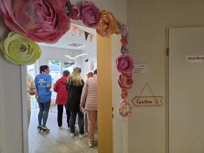 Menschen stehen in einem hellen Flur, dekoriert mit großen Papierblumen und Wimpelketten. Ein Schild mit der Aufschrift 