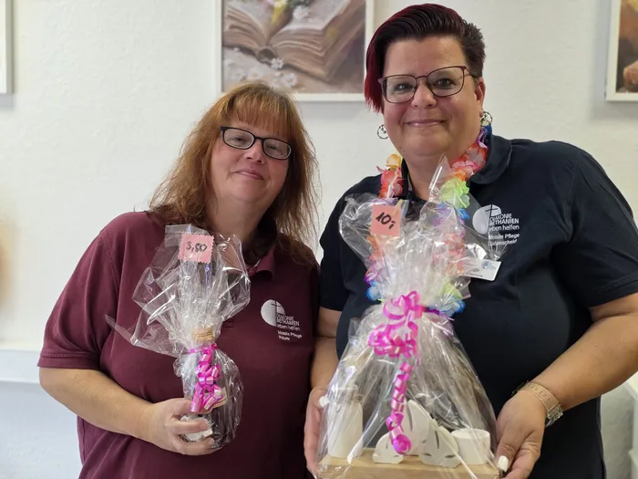 Zwei Frauen mit je einem kleinen, mit Schleifen versehenen Geschenk in der Hand stehen vor einer hellen Wand. Beide tragen Namensschilder mit dem Aufdruck 
