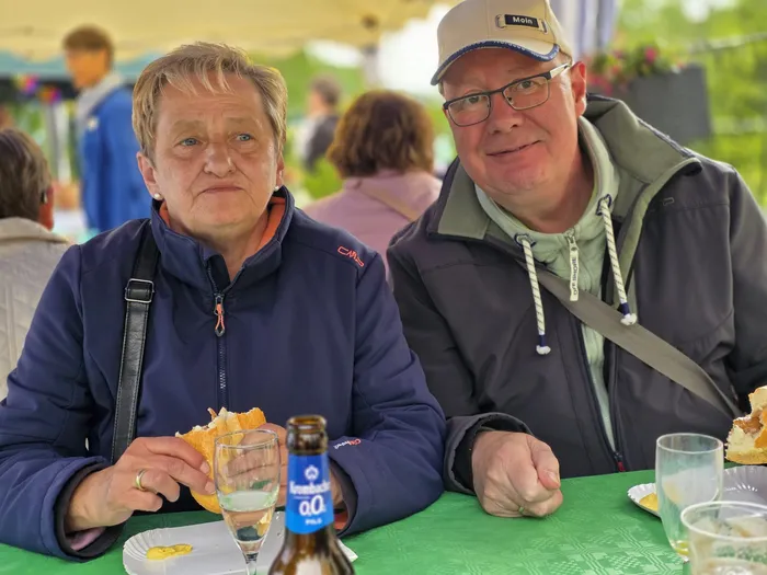 Zwei Personen sitzen an einem Tisch und essen Sandwiches. Im Hintergrund sind weitere Menschen und Festzeltplanen zu sehen.
