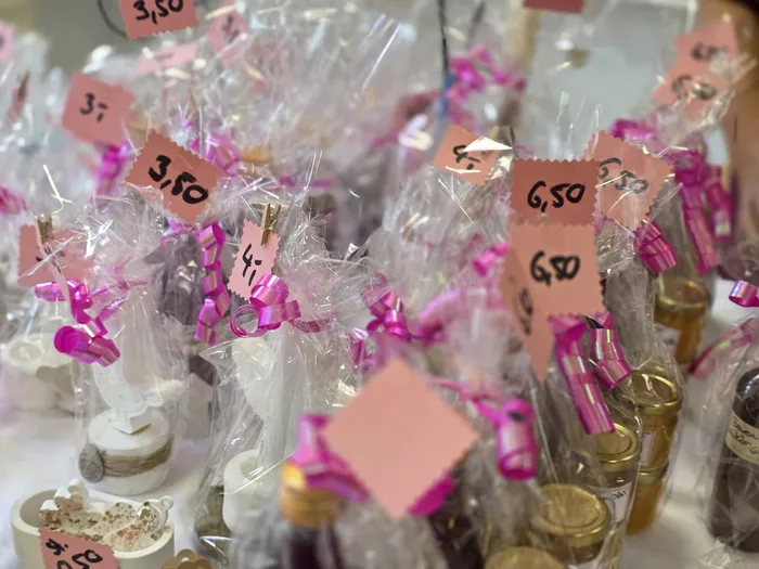 Verschiedene Süßigkeiten und kleine Geschenke, verpackt in durchsichtige Plastiktüten mit rosa Schleifen, stehen auf einem Tisch und sind mit Preisschildern versehen. Im Hintergrund sind weitere Artikel und Menschen zu erkennen.