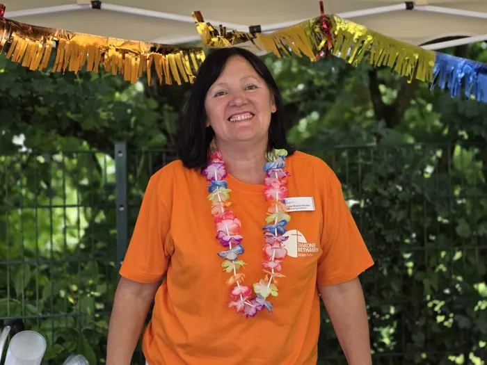 Eine Frau mit kurzen, braunen Haaren lächelt in die Kamera. Sie trägt ein orangefarbenes T-Shirt mit dem Logo der Diakonie Bethanien und eine bunte Blumenkette. Im Hintergrund ist ein festlich geschmückter Pavillon zu sehen.