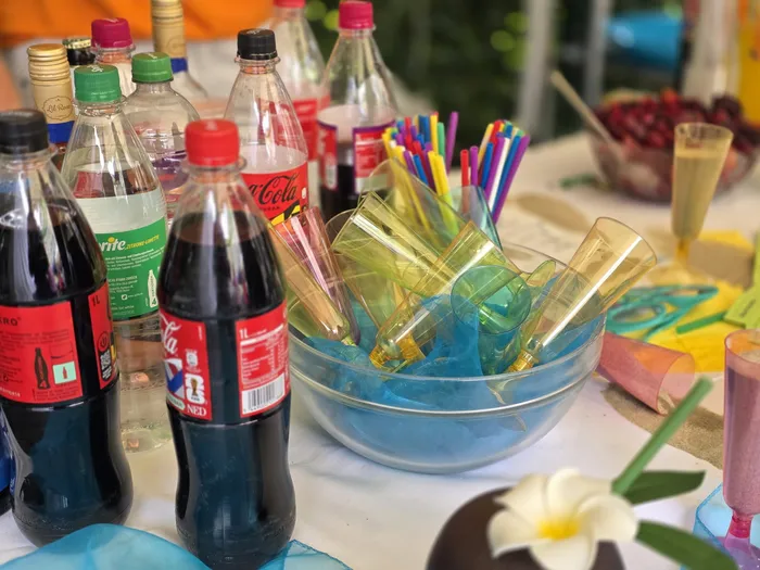 Getränke in Flaschen und Gläsern stehen auf einem mit einer blauen Tischdecke bedeckten Tisch, daneben bunte Strohhalme und eine Schale mit Eiswürfeln. Im Hintergrund sind weitere Gegenstände und grüne Pflanzen zu sehen.