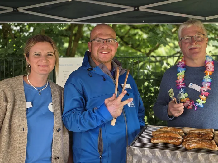 Drei Personen stehen hinter einem Grillwagen auf einem Sommerfest. Die Frau links und der Mann in der Mitte tragen Namensschilder und blaue Jacken, der Mann rechts eine Hawaii-Blumenkette und eine Brille. Auf dem Grill liegen Bratwürste.