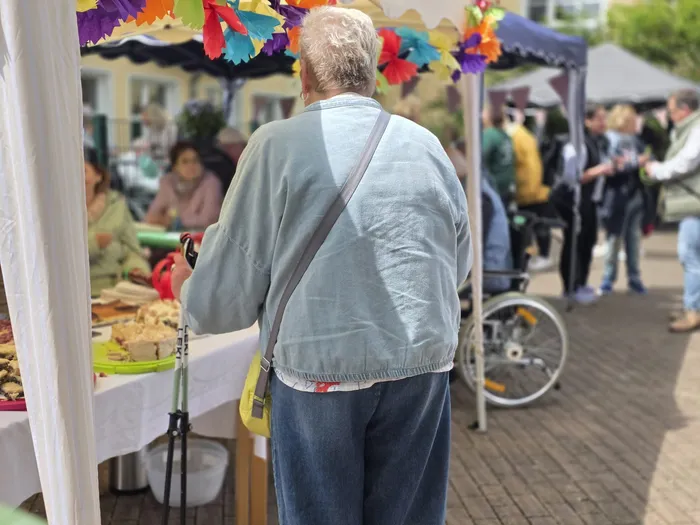 Ein älterer Mann mit grauem Haar steht mit dem Rücken zum Betrachter an einem Marktstand und betrachtet das Angebot. Im Hintergrund sind weitere Menschen und ein Rollstuhl zu sehen.