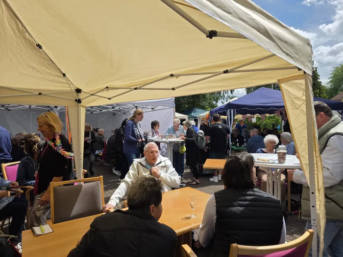 Menschen sitzen an Tischen unter einem gelben Zelt und essen und trinken. Im Hintergrund sind weitere Zelte und Besucher eines Sommerfestes zu sehen.