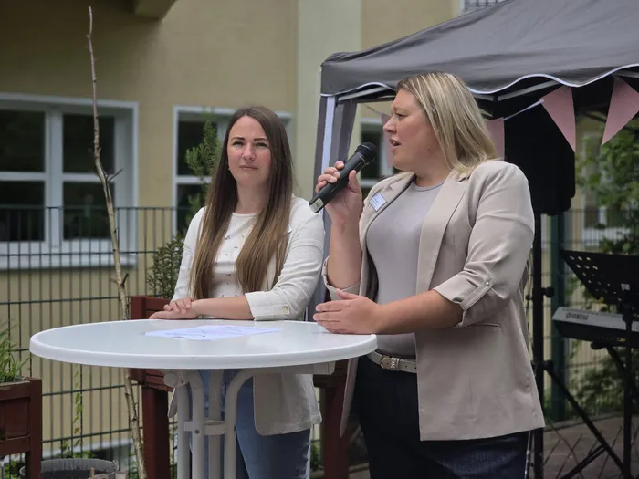 Zwei Frauen stehen an einem Tisch mit einem weißen Tischdecken. Die Frau rechts hält ein Mikrofon und spricht, während die Frau links ihr zuhört. Im Hintergrund steht ein Keyboard und ein Pavillon.