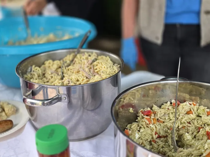 Zwei große Edelstahltöpfe gefüllt mit Nudelsalat stehen auf einem mit einer weißen Tischdecke bedeckten Tisch. Im Hintergrund sind Personen mit blauen Handschuhen zu sehen.