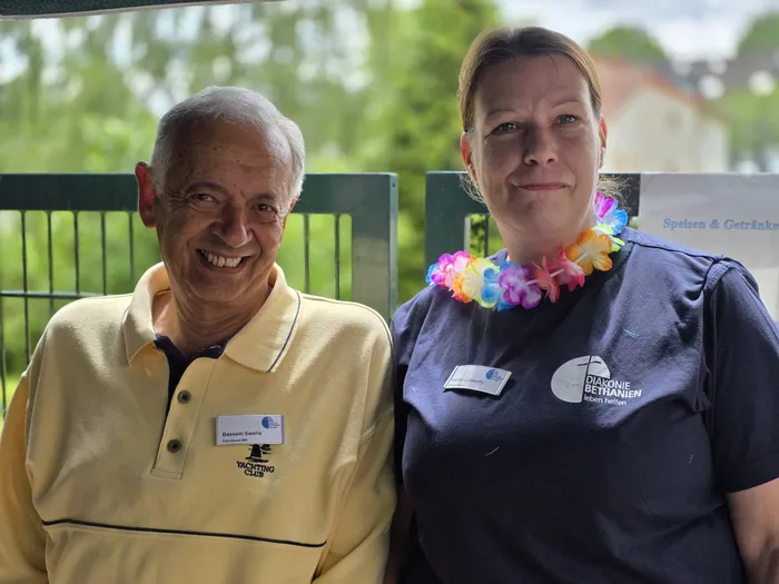Zwei Personen lächeln in die Kamera. Der Mann links trägt ein gelbes Poloshirt mit einem Yachtclub-Logo und einen Namensschild mit der Aufschrift „Bassem Sweha“. Die Frau rechts trägt ein blaues Hemd mit einem Anstecker der „Diakonie Bethanien“ und eine bunte Blumenkette.