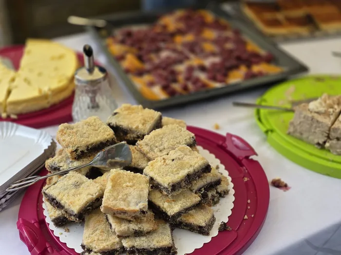 Ein roter Servierteller mit gestapelten, quadratischen Kuchenstücken im Vordergrund. Im Hintergrund sind weitere Kuchen und Torten auf einem Tisch zu sehen.