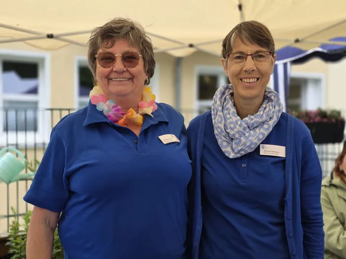 Zwei lächelnde Frauen mit Namensschildern stehen vor einem weißen Zelt. Sie tragen blaue Poloshirts und Accessoires, die zum Sommerfest passen.