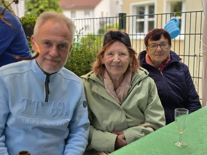 Drei ältere Menschen sitzen an einem Tisch im Freien und lächeln in die Kamera. Im Hintergrund ist ein Gebäude und Grünpflanzen zu sehen.