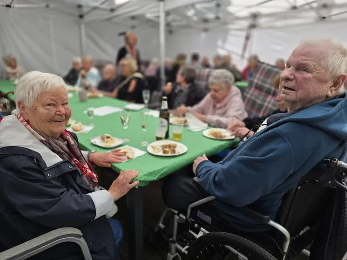 Zwei ältere Menschen sitzen an einem Tisch unter einem Zelt und unterhalten sich während eines Essens. Im Hintergrund sind weitere Menschen bei einem Sommerfest zu sehen.