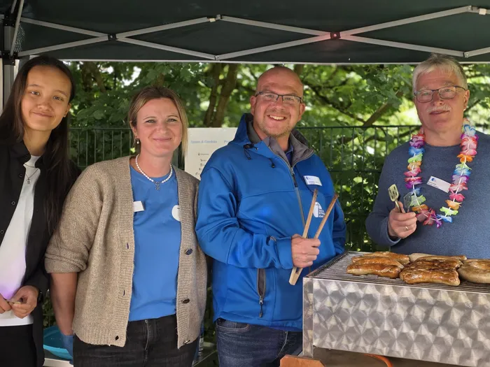 Vier Personen stehen vor einem Stand mit einem Sonnensegel. Ein Mann brät Würstchen auf einem Grill, während die anderen drei zusehen. Im Hintergrund sind Bäume zu sehen.