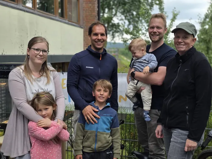 Eine Gruppe von Erwachsenen und Kindern steht lächelnd vor einem Gebäude mit einem grünen Hintergrund. Im Vordergrund hält ein Mann ein kleines Kind, während eine Frau neben ihm steht und ein weiteres Kind an der Hand führt.