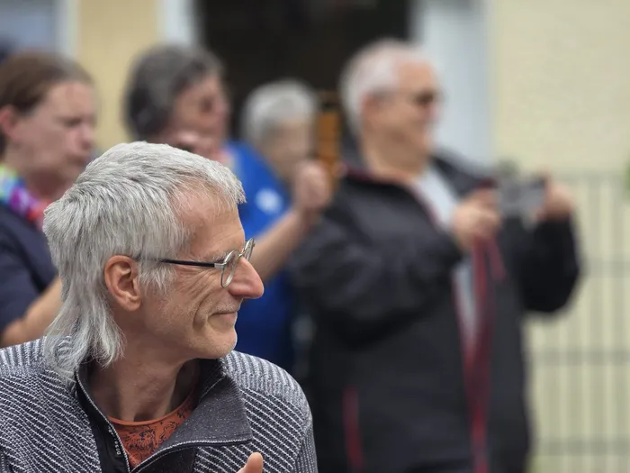Ein älterer Mann mit grauem Haar und Brille lächelt in die Kamera, im Hintergrund weitere Menschen bei einem Sommerfest.
