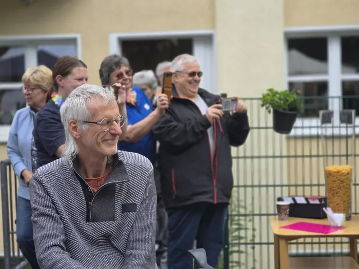 Ein lächelnder Mann mit grauen Haaren und Brille sitzt im Vordergrund. Im Hintergrund sind weitere Menschen bei einem Sommerfest zu sehen, einige fotografieren mit ihren Handys.