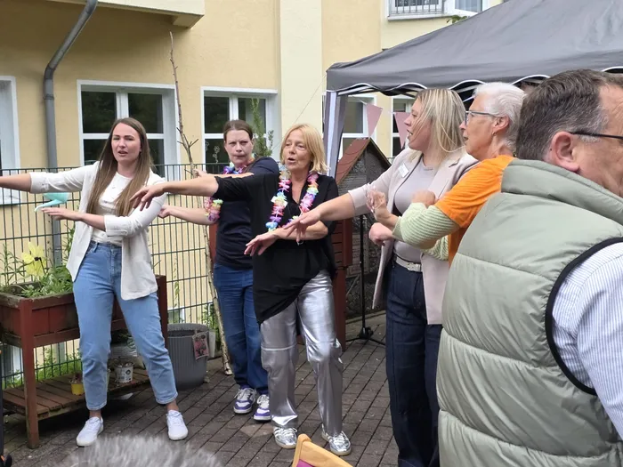 Eine Gruppe von sechs Personen steht im Freien und hält die Arme ausgestreckt, als ob sie eine Übung machen oder tanzen. Im Hintergrund ist ein Gebäude und ein Pavillon zu sehen.