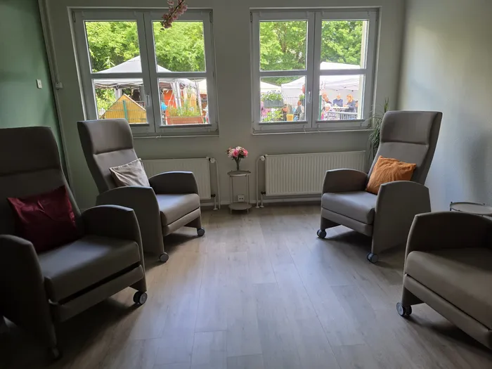 Vier bequeme Sessel stehen in einem hellen Raum mit großen Fenstern, die auf einen Garten mit bunten Zelten und Menschen blicken. Das Bild zeigt einen Aufenthaltsraum in der Tagespflege Bethanien während des Sommerfests.