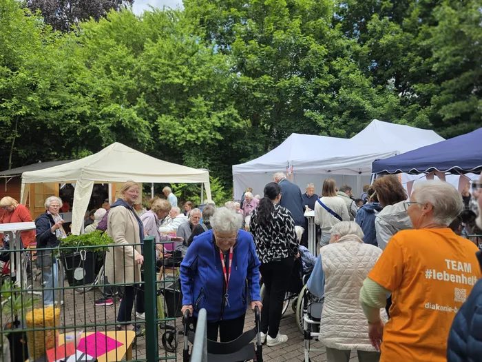 Menschen unterschiedlichen Alters sitzen und stehen auf einem Sommerfest unter weißen Zelten. Eine Frau mit Rollator geht im Vordergrund durch die Menge.