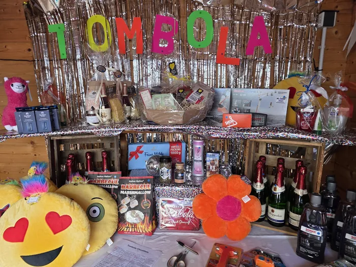 Ein Tisch ist voll mit Preisen für ein Tombola-Spiel, darunter Lebensmittel, Spirituosen, Spielzeug und Haushaltsartikel. Im Hintergrund ist ein Banner mit der Aufschrift 'TOMBOLA' zu sehen.
