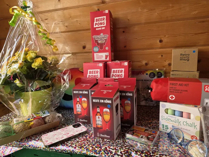 Mehrere Geschenkkörbe und -pakete stehen auf einem Holzboden. Darunter befinden sich Beer Pong Sets, Getränke, ein Erste-Hilfe-Set und Straßenmalkreide. Links sind rote Blumen zu sehen.