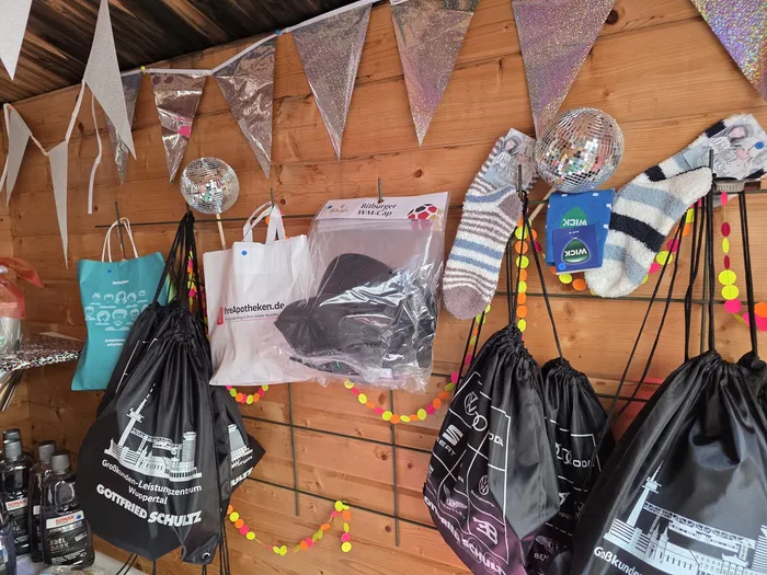 A variety of promotional items and giveaways hang on a wooden wall decorated with colorful pennants. Bags with logos, a disco ball, and other items suggest a festive event.