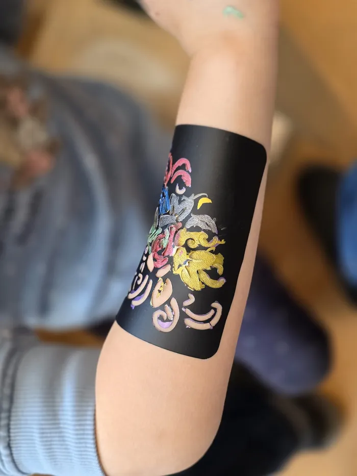 Ein Arm trägt ein temporäres Tattoo mit bunten floralen Mustern und einem kleinen Drachen auf schwarzem Hintergrund. Im Hintergrund ist ein Teil einer Person mit einem farbenfrohen Hemd zu sehen.