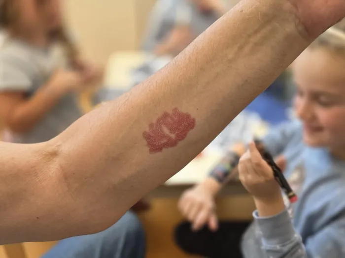 Ein Unterarm mit einem roten Lippenabdruck darauf. Im Hintergrund sind verschwommen Kinder und Erwachsene bei einer Veranstaltung zu sehen.