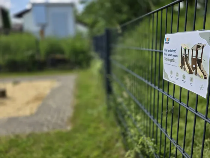 Ein Schild an einem Maschendrahtzaun kündigt an, dass hier bald ein neues Spielgerät entsteht. Im Hintergrund ist ein Park mit Bäumen und Büschen zu sehen.