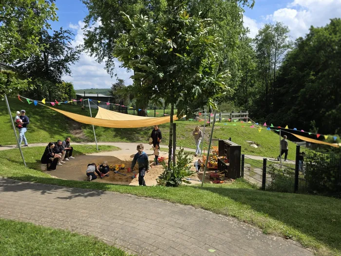 Kinder spielen im Sandkasten unter einem Sonnensegel, während Eltern und andere Besucher am Rande zusehen. Im Hintergrund sind Bäume und eine Girlande mit bunten Flaggen zu sehen.