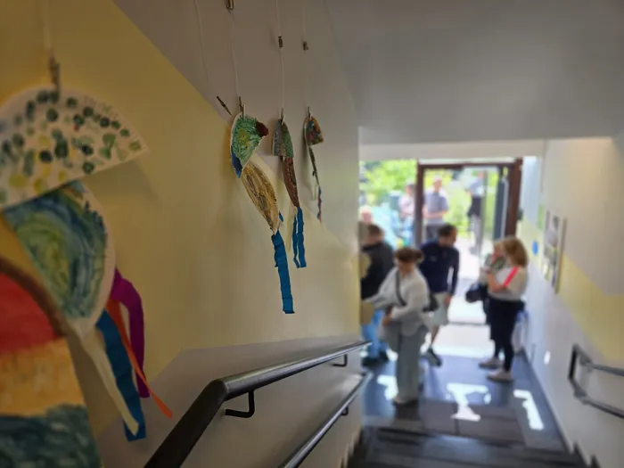 Kinderarbeiten, bunte Drachen aus Papier, hängen an der hellen Wand einer Treppe. Im Hintergrund sind Besucher des Tages der offenen Tür der KiTa Wunderland zu sehen.