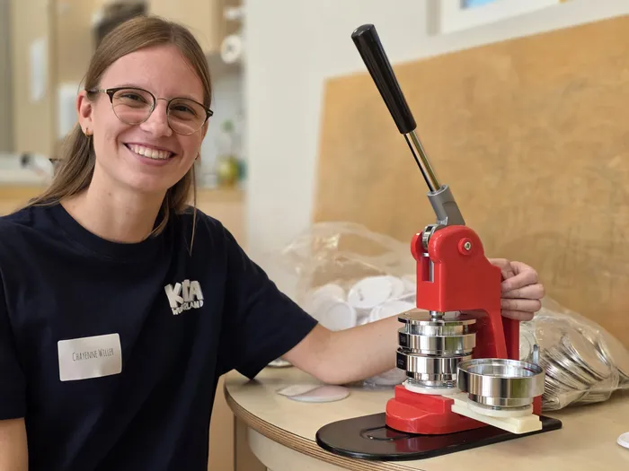 Junge Frau mit Brille lächelt in die Kamera, während sie eine rote Buttonmaschine bedient. Vor ihr liegen Buttons und ein Namensschild mit der Aufschrift „Chayenne Willer“.