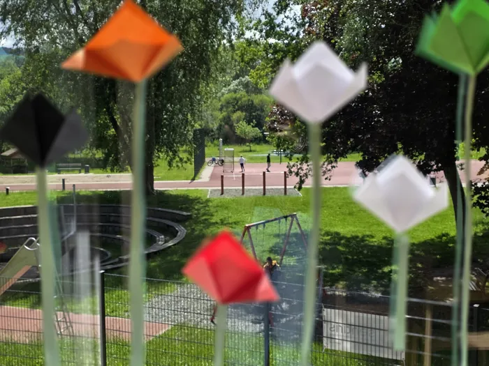 Ein Spielplatz mit Schaukeln und einem Karussell ist durch bunte, origamiartige Blumen im Vordergrund zu sehen. Im Hintergrund sind Menschen und Fahrräder auf einem Weg zu erkennen.
