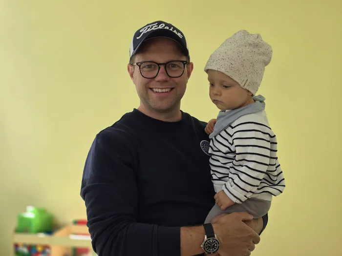 Ein Mann mit Brille und Baseballkappe hält ein Baby in den Armen. Beide schauen in die Kamera. Im Hintergrund ist ein Raum mit Spielzeug zu sehen.