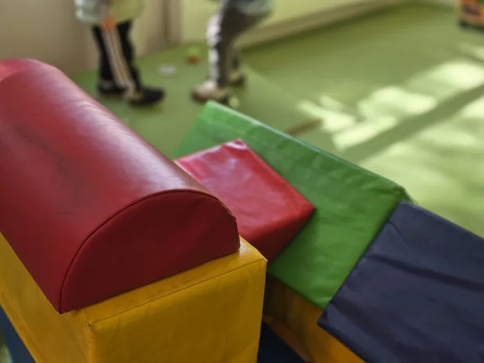 Bunte Schaumstoffbausteine in Rot, Gelb, Grün und Blau stehen auf einer grünen Matte. Im Hintergrund sind die Beine von Personen zu sehen.