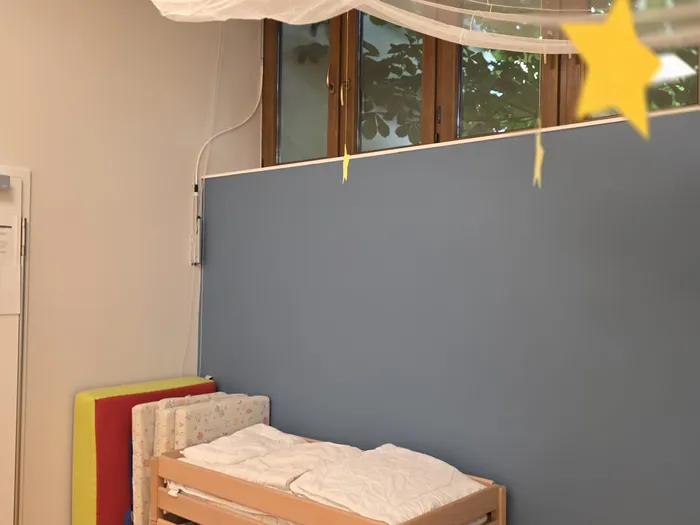 Ein Kinderbett mit weißem Bettzeug steht in einem Raum mit hellblauen Wänden. Über dem Bett hängen gelbe Sterne und ein weißer Stoffabhang vor einem Fenster.