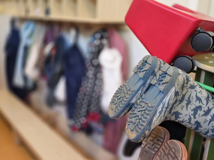 Ein Paar schmutziger, blauer Kinderschuhe mit Dinosaurier-Motiv hängt an einem roten Kinderstuhl. Im Hintergrund sind Regale mit Kinderkleidung zu sehen.