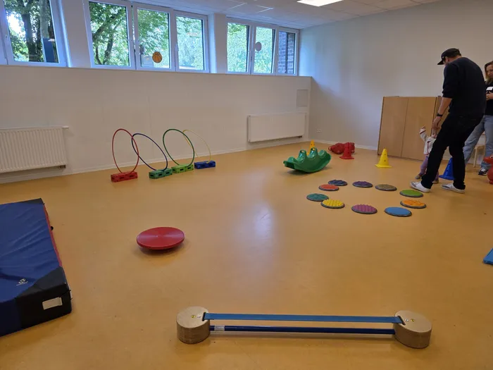 Ein großer, heller Raum mit Spielgeräten für Kinder, darunter Bälle, Reifen, eine Wippe und Balance Boards. Im Hintergrund sind die Beine von Personen zu sehen, die den Tag der offenen Tür der KiTa Wunderland besuchen.