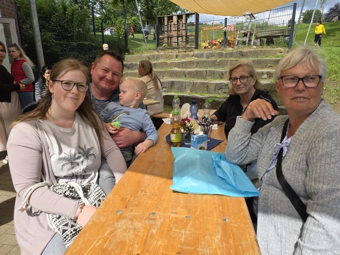 Eine Gruppe von Erwachsenen und ein Kleinkind sitzen an einem Holztisch im Freien, umgeben von weiteren Menschen und einem Spielplatz im Hintergrund. Sie scheinen sich bei einem Treffen der Kita Wunderland zu unterhalten.