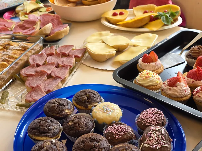 Ein Tisch ist mit verschiedenen Speisen wie Muffins, Wurst, Käse, Obst und Gebäck gedeckt. Im Hintergrund sind Personen zu erkennen, die an dem Tag der offenen Tür der KiTa Wunderland teilnehmen.