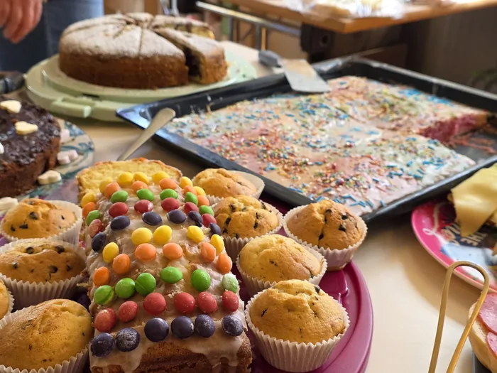 Verschiedene selbstgebackene Kuchen und Muffins stehen auf einem Tisch, dekoriert mit bunten Streuseln und Süßigkeiten. Im Hintergrund sind weitere Speisen und Menschen zu erkennen, die an einem Tag der offenen Tür der KiTa Wunderland teilnehmen.