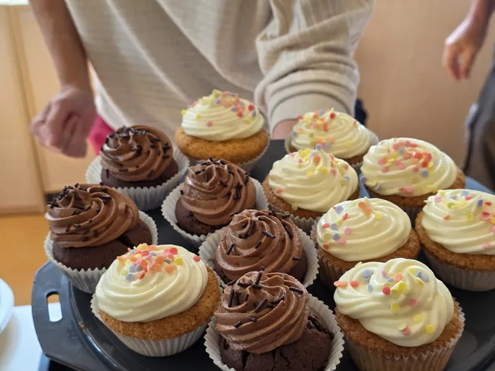 Ein Tablett mit gemischten Cupcakes, einige mit Schokoladen-, andere mit Vanillecreme und bunten Streuseln, steht im Fokus. Im Hintergrund ist ein Teil einer Person zu sehen, die möglicherweise die Cupcakes anbietet.