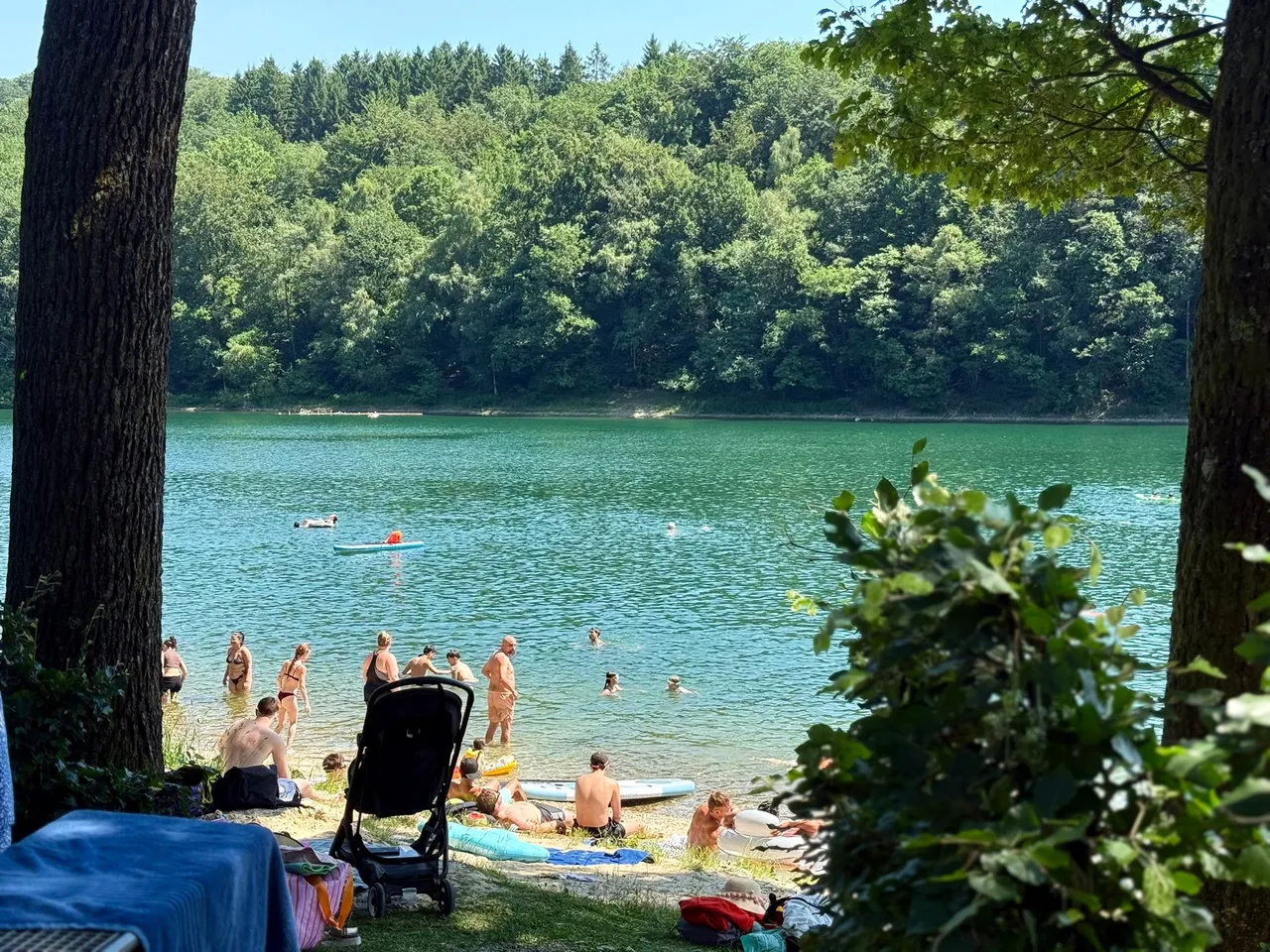 Menschen schwimmen und liegen an einem sonnigen Tag an der Glörtalsperre. Am Ufer liegen Handtücher, ein Kinderwagen und persönliche Gegenstände.