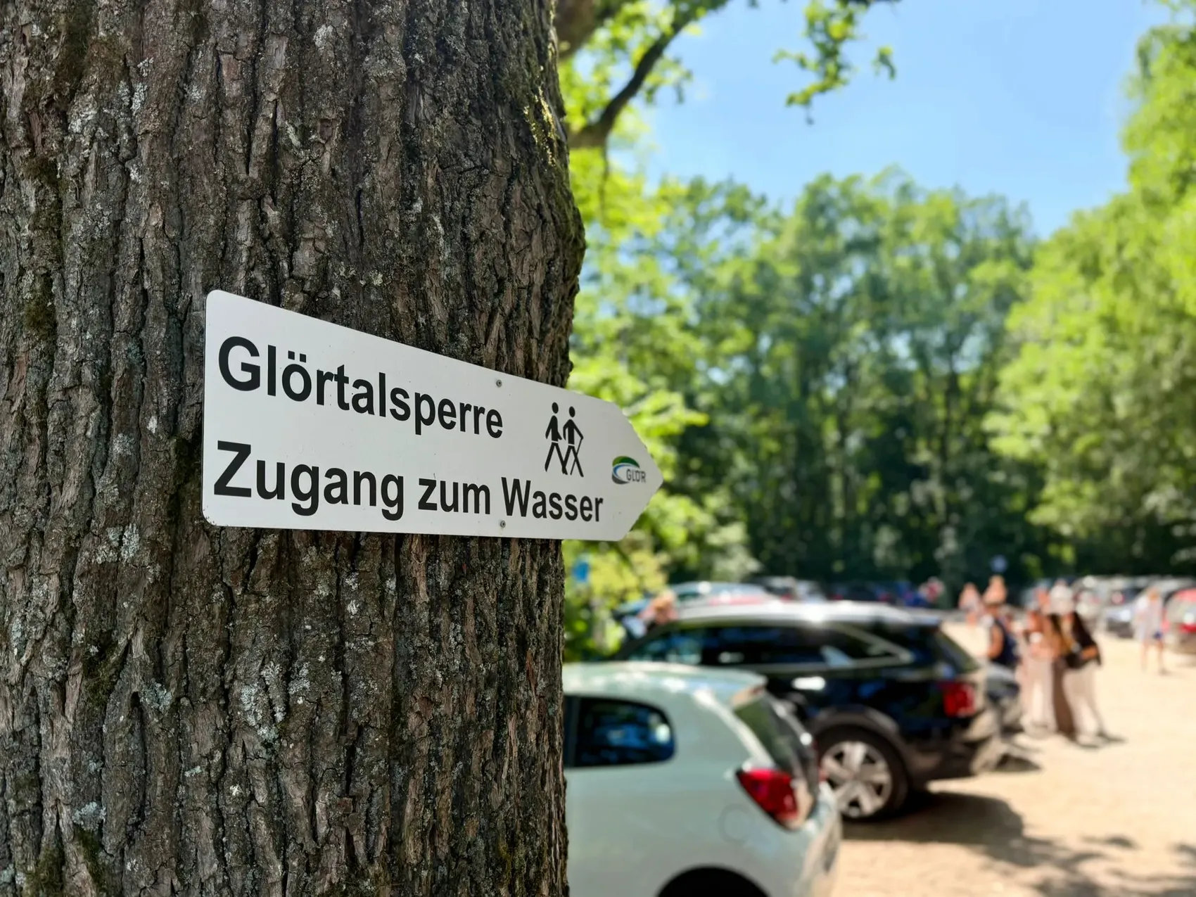 Ein Schild an einem Baum weist auf die „Glörtalsperre“ und den „Zugang zum Wasser“ hin. Im Hintergrund sind Autos und Menschen zu sehen.