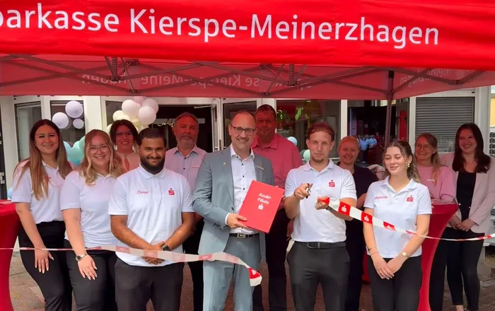Gruppe von Mitarbeitern der Sparkasse Kierspe-Meinerzhagen vor dem Eingang einer Filiale, schneiden ein rotes Band durch.