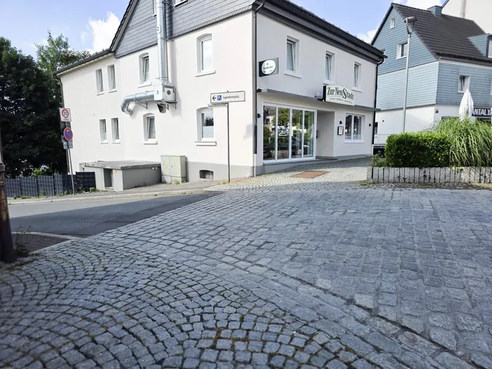Ein helles Gebäude mit dem Schild „Zur Neuen Stadt“ steht an einer Kreuzung mit einem Kopfsteinpflasterweg. Ein Hinweisschild zeigt den Weg zu einem Parkplatz.