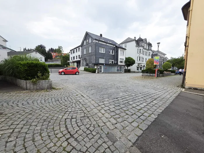 Ein Platz mit Kopfsteinpflaster in einer Wohngegend. Im Hintergrund stehen mehrere Häuser, darunter ein Gebäude mit dunkler Fassade und einem roten Auto davor. Der Himmel ist bewölkt.