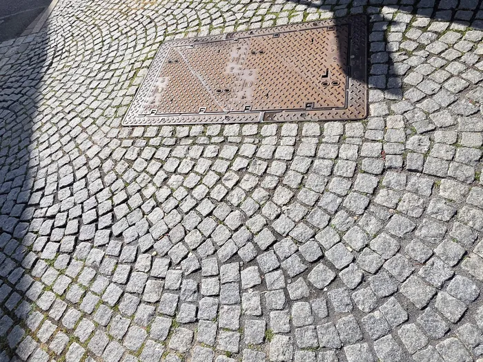 Ein quadratischer Gullydeckel aus Metall ist in ein Pflaster aus kleinen grauen Steinen eingelassen. Der Deckel zeigt ein Muster aus kleinen Löchern und hat eine Markierung an einer Ecke.